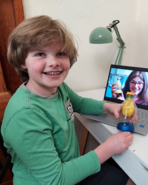 Student smiling and holding a lung model they created that matches the model Dr. Robin is holding up in a recorded lesson playing on the computer screen behind the student